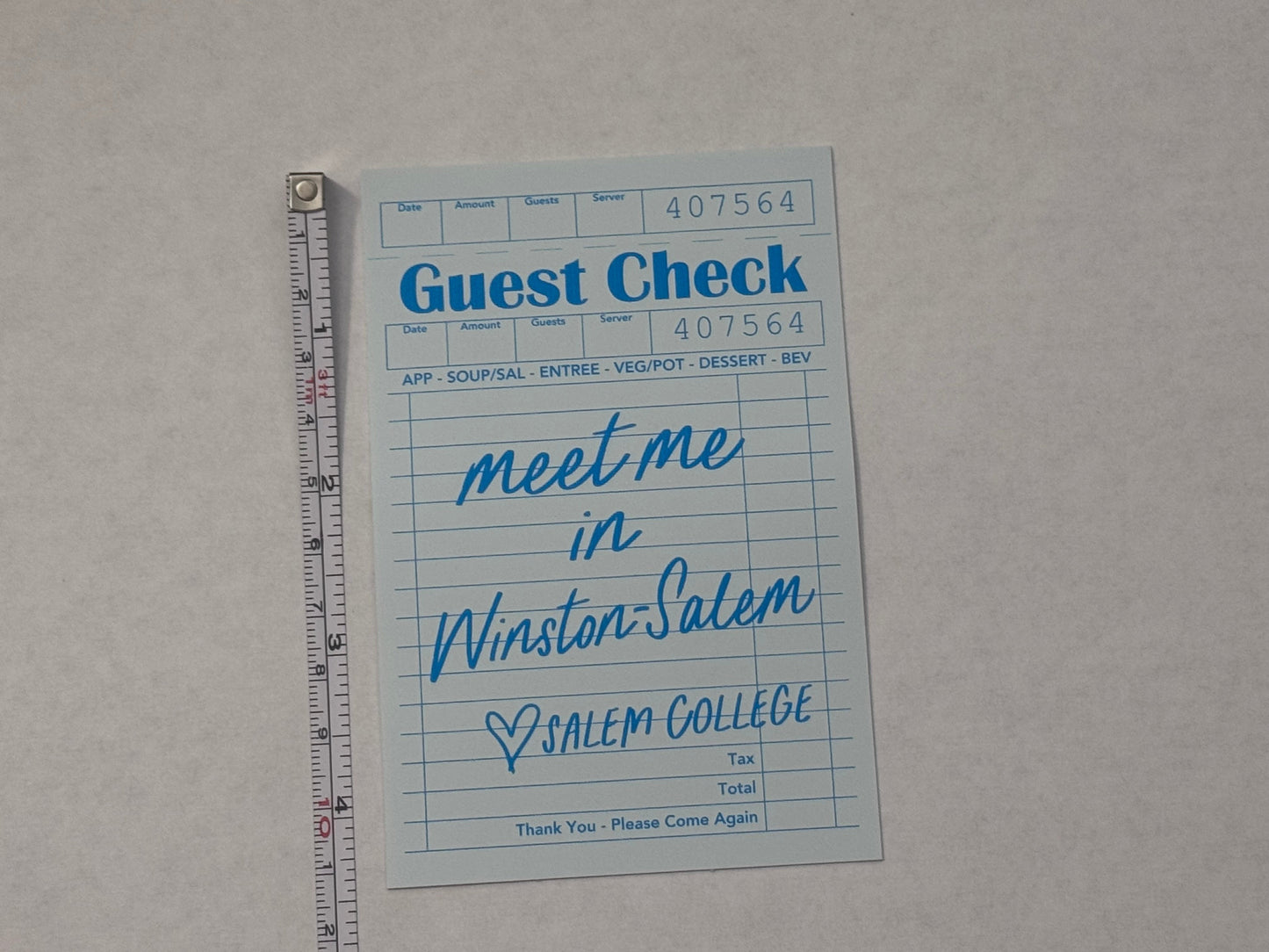 A light blue guest check card with handwritten text reading 'Meet me in Winston-Salem, NC' placed on a light gray surface with a metal ruler above it for scale.