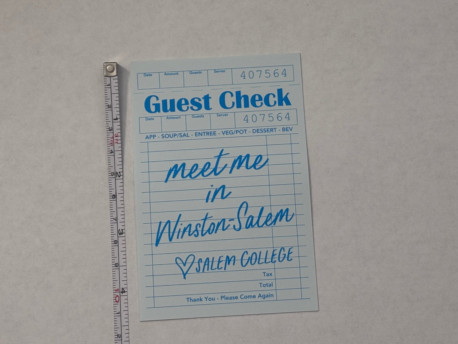 A light blue guest check card with handwritten text reading 'Meet me in Winston-Salem, NC' placed on a light gray surface with a metal ruler above it for scale.