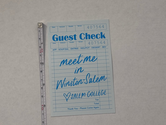 A light blue guest check card with handwritten text reading 'Meet me in Winston-Salem, NC' placed on a light gray surface with a metal ruler above it for scale.