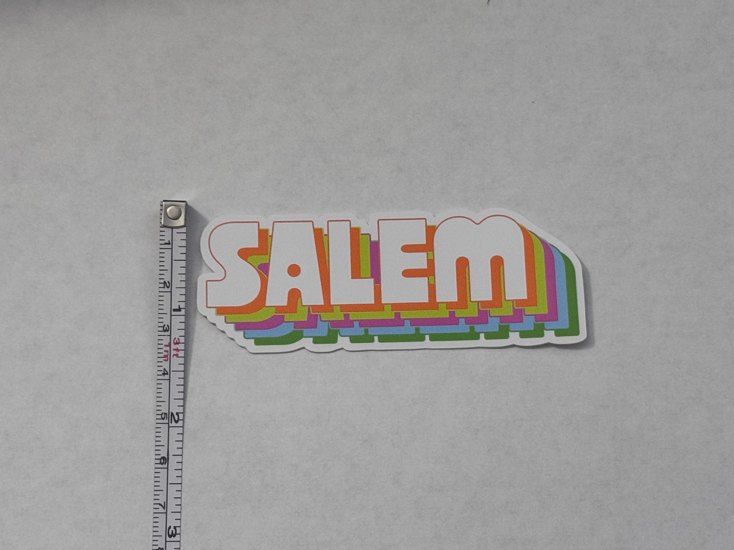 A die-cut vinyl sticker featuring the word 'SALEM' in a retro layered rainbow design, shown next to a metal tape measure on a light gray background.