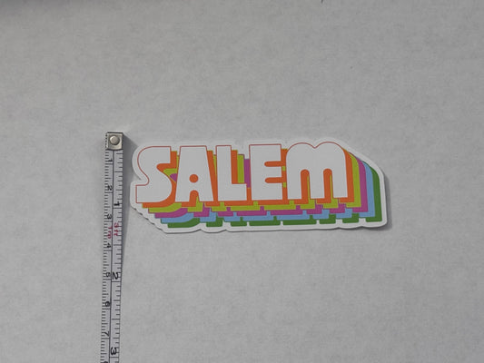 A die-cut vinyl sticker featuring the word 'SALEM' in a retro layered rainbow design, shown next to a metal tape measure on a light gray background.